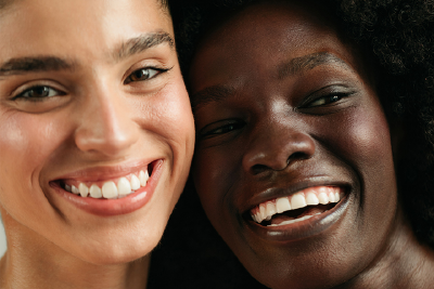 "Close-up of two women with radiant skin smiling and leaning close to each other, showcasing diverse beauty and natural joy."